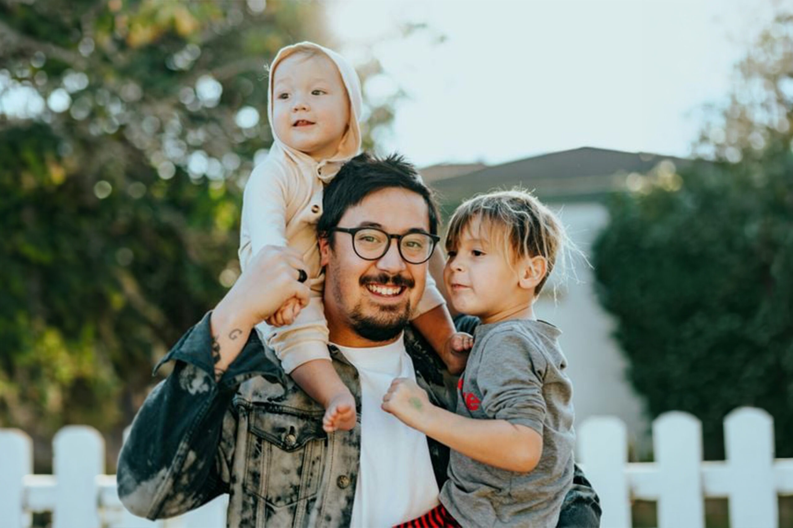 A smiling man holds two young children, one on his shoulders and another in his arms, against a bright, outdoor backdrop.
