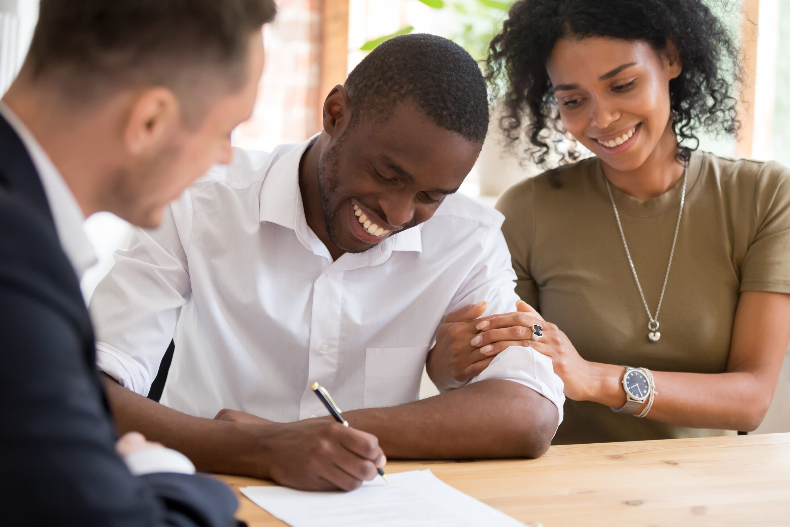 A man smiles while signing a document, supported by a woman beside him. Another man looks on, creating a supportive atmosphere for a transaction.