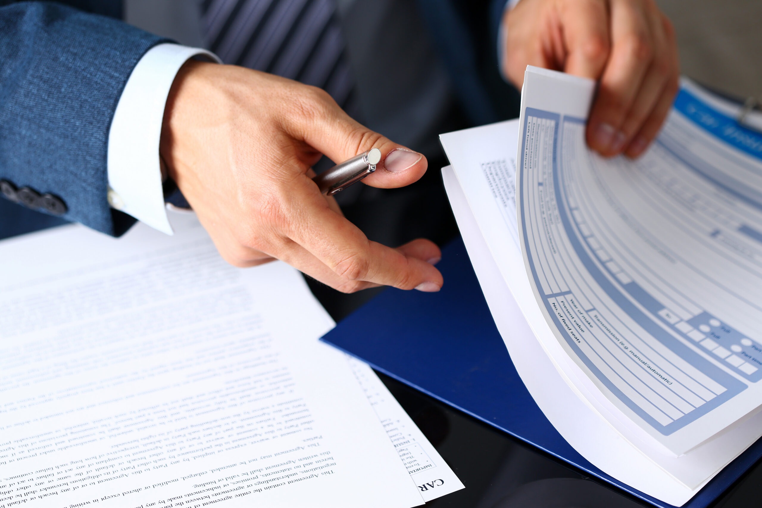 A professional hand holds a pen while sorting through paperwork, indicating a focus on organization and detail in a business setting.